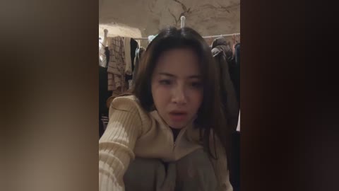 Media: Video of an Asian woman with long, straight dark hair, wearing a beige sweater, looking surprised, with a beige lamp and hanging clothes in the background.