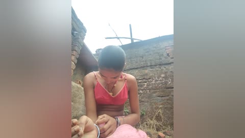 Media: Video of a young Indian woman with dark skin and straight black hair tied back, wearing a pink tank top and bracelets, kneeling in a rural setting, feeding a baby.