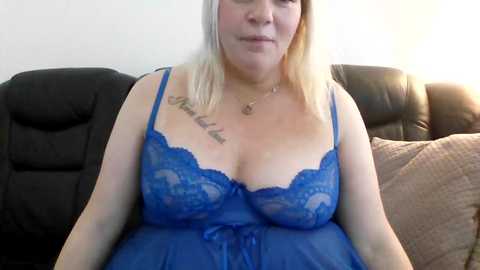 A video of a blonde woman in a sheer blue lace nightgown with a tattoo on her chest, sitting on a black couch in a dimly lit room.