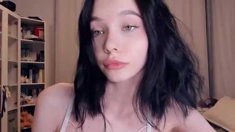 Video of a young woman with pale skin, straight black hair, and light makeup, wearing a white camisole. She stands in a cluttered room with bookshelves and a beige curtain in the background.