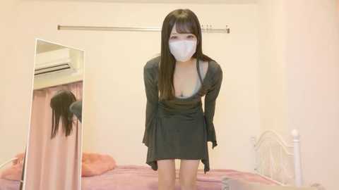 A video of an Asian woman in a black dress with a plunging neckline, wearing a white face mask, standing in a beige room with a mirror, bed, and air conditioner.