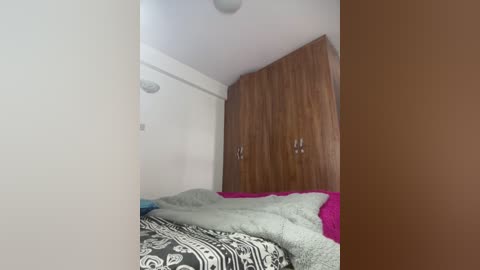A video of a small, dimly lit bedroom with white walls, a bed covered in a black and white patterned duvet, and a wooden wardrobe with silver handles.