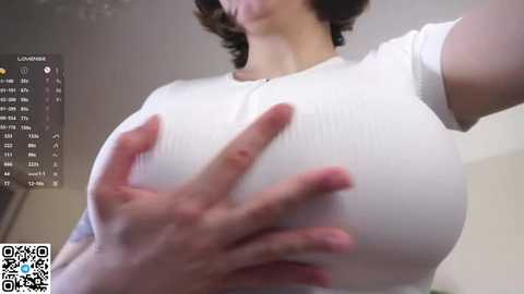 Video of a woman with large breasts, wearing a white ribbed top, hands cupping her breasts. Background shows a blurred indoor setting with a calendar and text.