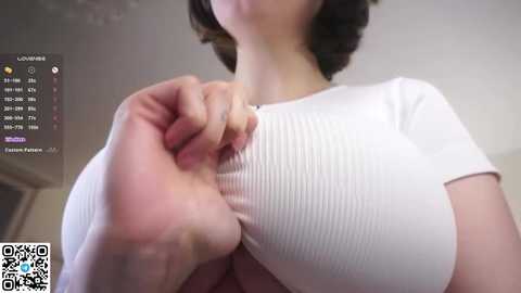 Video of a light-skinned woman with short brown hair, wearing a tight white ribbed top that accentuates her large breasts. Background shows a digital screen with text and icons.