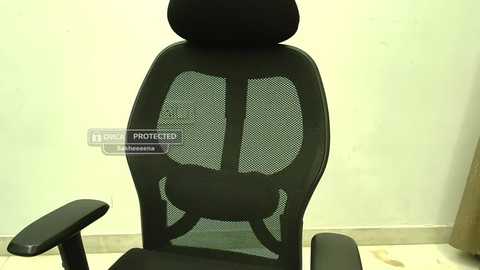 Video of a black mesh office chair with a high backrest and armrests, positioned against a plain light green wall. The chair's seat is partially visible, showing its mesh material.