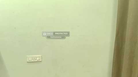 A video of a beige wall with a white rectangular electrical outlet and a small rectangular sign labeled \"PROTECTED\" and \"EMERGENCY EXIT\" in white text. The background is a beige curtain.