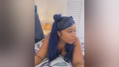 Video of a dark-skinned woman with blue hair in buns, wearing a black top, lying on a bed with white and blue patterned sheets, in a softly lit bedroom with a bedside lamp and white shutters.