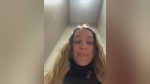 Media: A video of a young Caucasian woman with long, wavy blonde hair and a black turtleneck sweater, making a silly face with her tongue out. She is standing in a narrow, dimly lit hallway with beige walls and tiled floor.