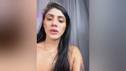 A close-up video of a young woman with long, straight black hair, light skin, and medium-sized breasts, wearing a gold necklace. Her expression is neutral, and she has a tattoo on her right arm.