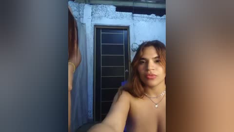 Video of a young, light-skinned woman with shoulder-length brown hair, topless, standing in a dimly lit, urban alley with a blue door in the background.