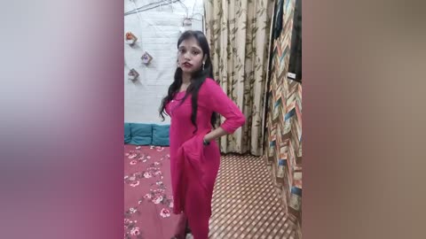 Video of a young woman in a pink salwar kameez standing in a small, cluttered room with floral-patterned curtains and a carpet.