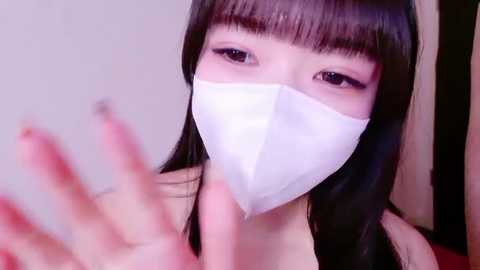 A video of an Asian woman with long black hair, wearing a white medical mask, holding up her hands in a playful gesture. The background is a blurred, light-colored room.