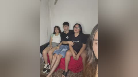 A video of four young people, three women and one man, sitting on a couch in a dimly lit room. They wear casual clothing, including a white t-shirt, denim shorts, and a black t-shirt. The background is plain and unadorned.