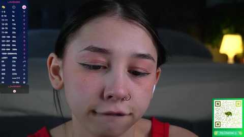 Media: A young Asian woman with dark hair, a septum piercing, and a red shirt, looking contemplative, against a dimly lit background with a green QR code overlay.