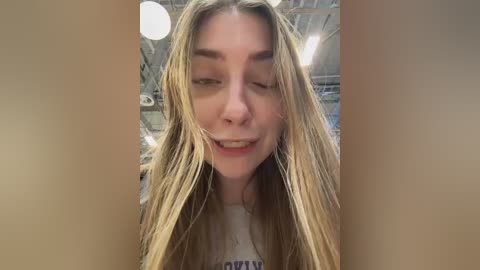Media: Video of a young Caucasian woman with long, wet, blonde hair, wearing a gray t-shirt, smiling and closing her eyes. The background shows a dimly lit, industrial room with visible pipes and ceiling lights.
