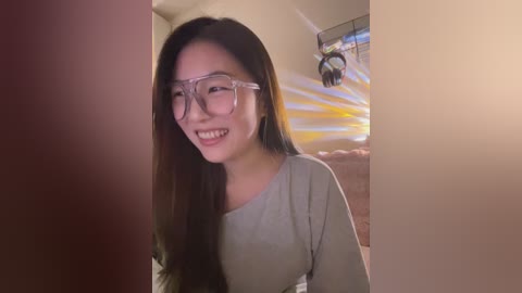 Video of a smiling Asian woman with long black hair, wearing clear glasses and a light grey top, standing in a dimly lit room with a sunburst wall hanging in the background.