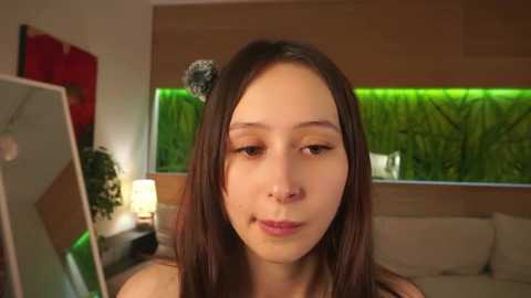 Video of a young, light-skinned woman with straight, shoulder-length brown hair, wearing a black top, staring into a mirror in a modern living room with green wall art and a sofa.