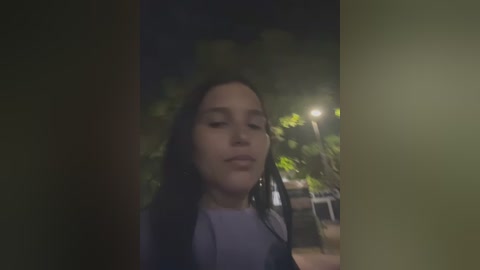 A video of a young woman with long black hair, closed eyes, and a neutral expression, standing in a dimly lit outdoor setting at night, surrounded by trees and a streetlamp.