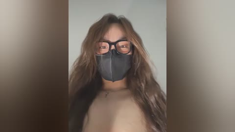 Media: Video of a woman with long brown hair, wearing black glasses and a black face mask, and a black top, against a neutral background.