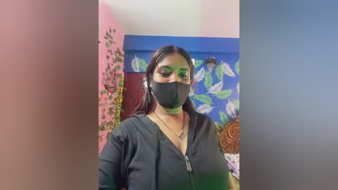 Video of a South Asian woman with medium skin tone, wearing a black face mask and black top, standing in a room with a blue feather patterned wall and pink wall, with greenish tint at the edges.