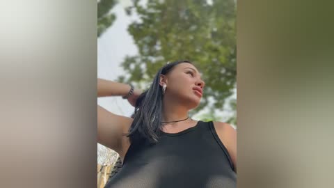 A video of a young woman with medium brown skin, straight black hair, and a slim physique, wearing a black tank top, standing outdoors with blurred green foliage and sunlight filtering through.