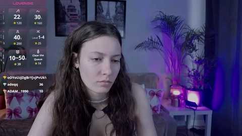 Video of a young woman with long, wavy brown hair and pale skin, wearing a black top, sitting on a couch in a dimly lit room with purple lighting, framed by a screen with various social media statistics.