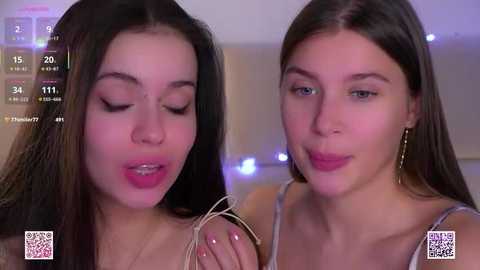 Video of two young women with straight, dark hair and light skin, wearing earrings, smiling. Background includes a blurred indoor setting with purple lighting and QR codes in the corners.