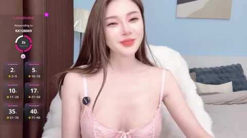 Video of a fair-skinned, slender woman with long brown hair, wearing a pink lace bra, smiling. Background features a white couch, blue wall, and a partially visible framed picture.