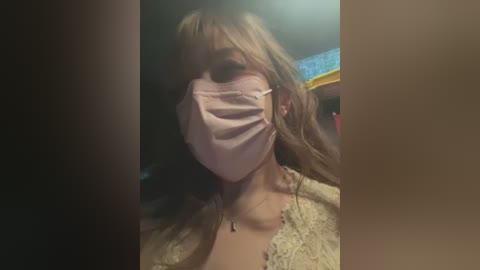 Video of a woman with long blonde hair, wearing a pink face mask, a white lace top, and a necklace, indoors with a blurred background.