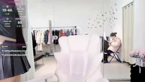 Video of a chic, modern clothing store with white walls and a mirror, showcasing a woman in a black skirt and a white chair, with a pink jacket on a hanger.