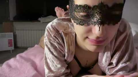 Video of a woman with light skin, wearing a black lace masquerade mask and a pink velvet robe, lying on a pink bedspread. Background includes a white radiator and a dark TV.