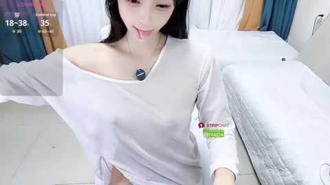 A video of an East Asian woman with long black hair, wearing a white off-shoulder sweater and sticking out her tongue, in a hospital room with white sheets and a teal curtain.