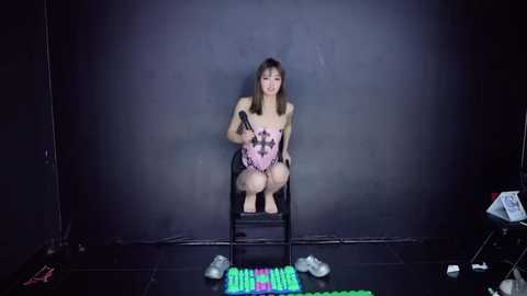 A video of an Asian woman with shoulder-length brown hair, dressed in a pink bodysuit, sitting on a black stool, holding a microphone, against a dark backdrop, with a colorful keyboard on the floor.