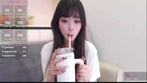 Media: Video of an Asian woman with long black hair, wearing a white t-shirt, drinking a milkshake through a straw, seated indoors on a gray sofa.
