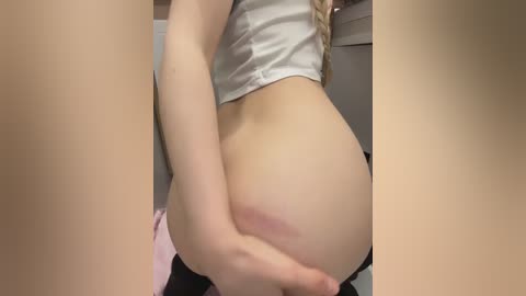 A close-up video of a woman with fair skin and a white crop top, bending over to reveal her bare buttocks. She has long blonde hair and is in a room with beige walls and a pink garment in the background.