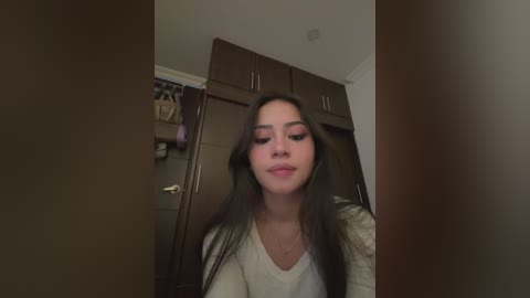 Media: Video of a young woman with long, straight black hair, fair skin, and a slight smile, wearing a light gray top. She stands in a dimly lit room with dark wooden wardrobes and a hanging pink item in the background.