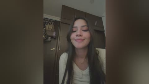 Media: A video of a young woman with long black hair, fair skin, and a slight smile, standing in a dimly lit, cluttered room with brown cabinets and hanging coats.