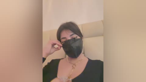 Media: A video of a woman with medium skin tone, wearing a black face mask, adjusting her glasses while seated indoors. She has a black top and a gold necklace. The background is beige.