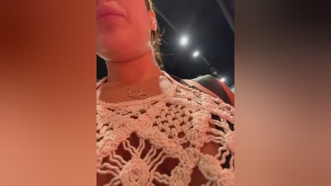 Video of a woman's upper body, partially obscured by a blurred, dark figure. She wears a beige, crocheted top with intricate patterns. Her skin is light, and she has a delicate necklace. The background shows warm, dim lighting and a blurred stage.