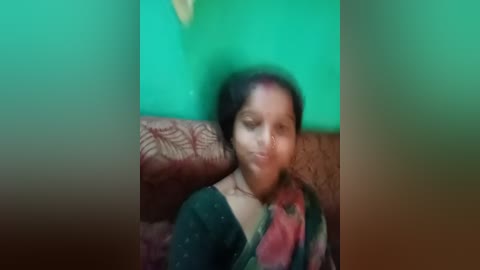 A video of a young South Asian woman with medium brown skin, wearing a dark green sari with floral patterns, sitting on a red cushioned sofa against a green wall. Her hair is dark and curly.
