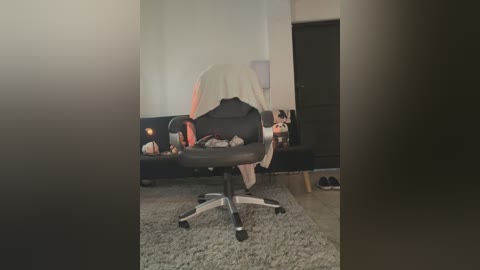 A video of a dark office chair with a white towel draped over it, positioned in a dimly lit room with a beige carpet, black couch, and wooden door.