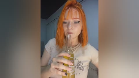 Video of a fair-skinned woman with bright orange hair and bangs, wearing a beige t-shirt, drinking from a glass bottle, in a dimly lit room with white walls and a dark ceiling.