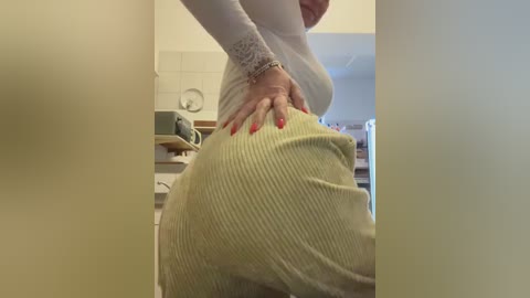 Video of a person with fair skin, wearing a white long-sleeve top and yellow ribbed skirt, pressing hands on hips, in a dimly lit kitchen with white tiles and wooden shelves.