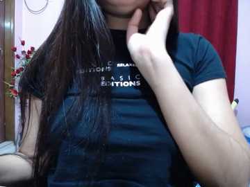 Video of a woman with long, dark hair, wearing a black t-shirt with white text, sitting indoors. She touches her face with her right hand. Red curtains and floral arrangement in the background.
