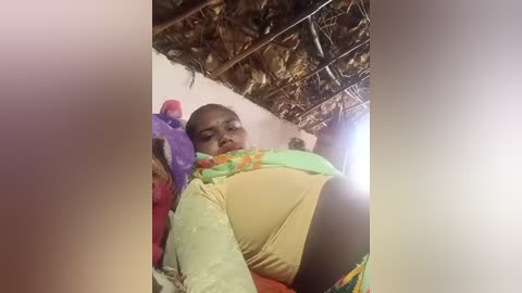 Video of an Indian woman with dark skin, wearing a colorful saree, sitting on a traditional mat inside a rustic hut with a thatched roof.