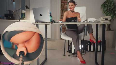 A professional woman with light skin, dark hair, and a black choker sits at a desk in a modern office, wearing a black strapless top and gray pants.