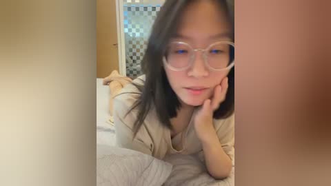 Media: Video of an Asian woman with long black hair, wearing oversized glasses, resting on her elbow, lying on a bed with gray sheets in a cozy, dimly lit room.
