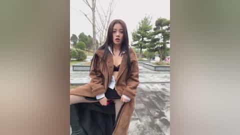 Video of a young Asian woman with long black hair, wearing a brown fur coat over a white shirt and black skirt, posing seductively on a stone bench in a garden.