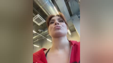 A video of a young woman with light skin and short auburn hair, wearing a red jacket, standing in an industrial corridor with metallic pipes and an air conditioning unit overhead.