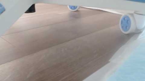 A video shows a beige wooden floor with two blue paint rollers being rolled horizontally, creating a smooth, even texture. The background is a light blue wall.